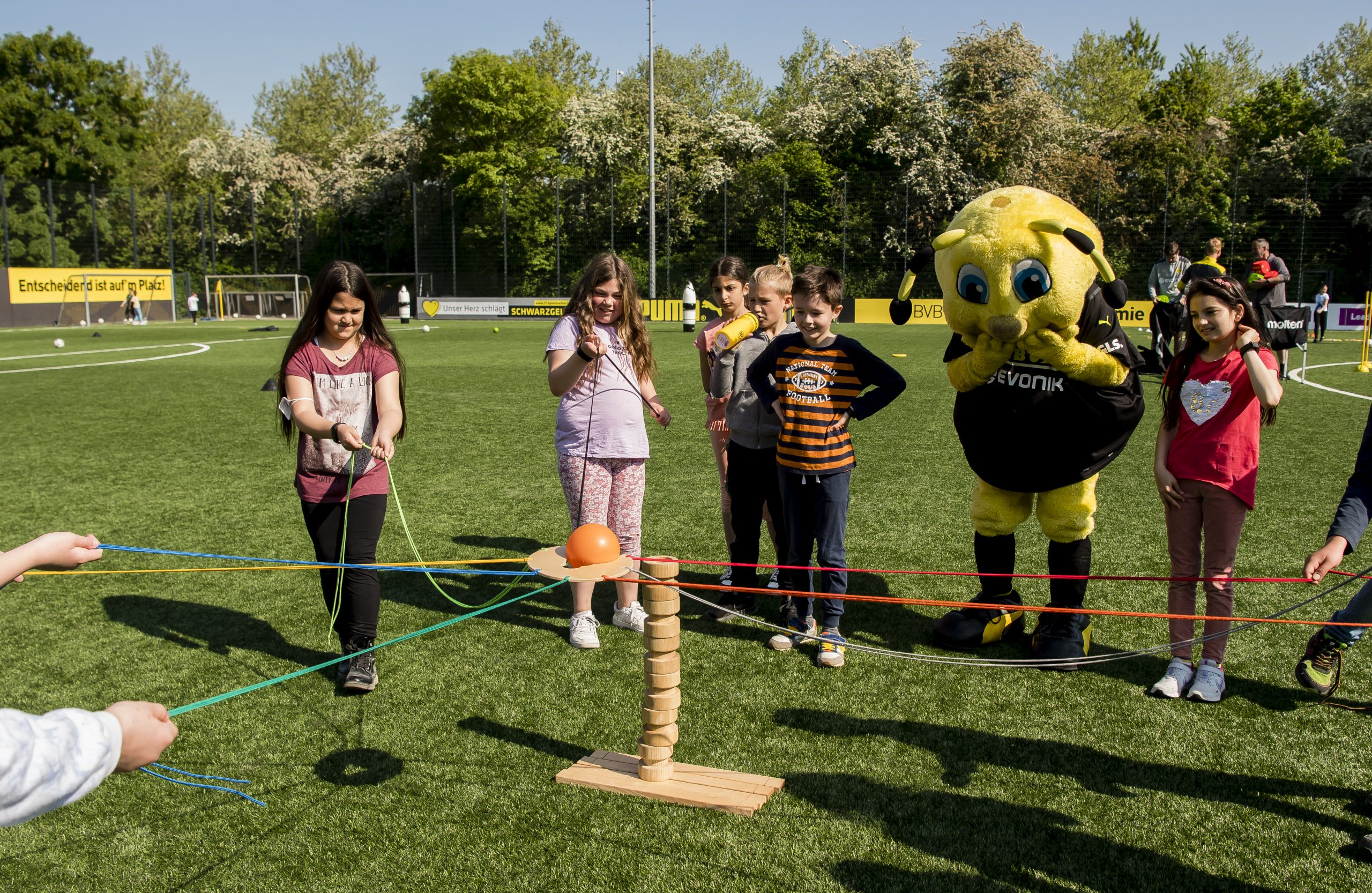 Drei silberne Wanderpokale für die Altersklassen U10, U12 und U14 stehen auf einer erhöhten Fläche vor einem Fußballfeld. Im Hintergrund sind spielende Kinder und Zuschauer zu sehen. Zwischen den Pokalen steht ein Schild mit der Aufschrift "TOP SCORER 18" und einer stilisierten Fußballspieler-Silhouette.