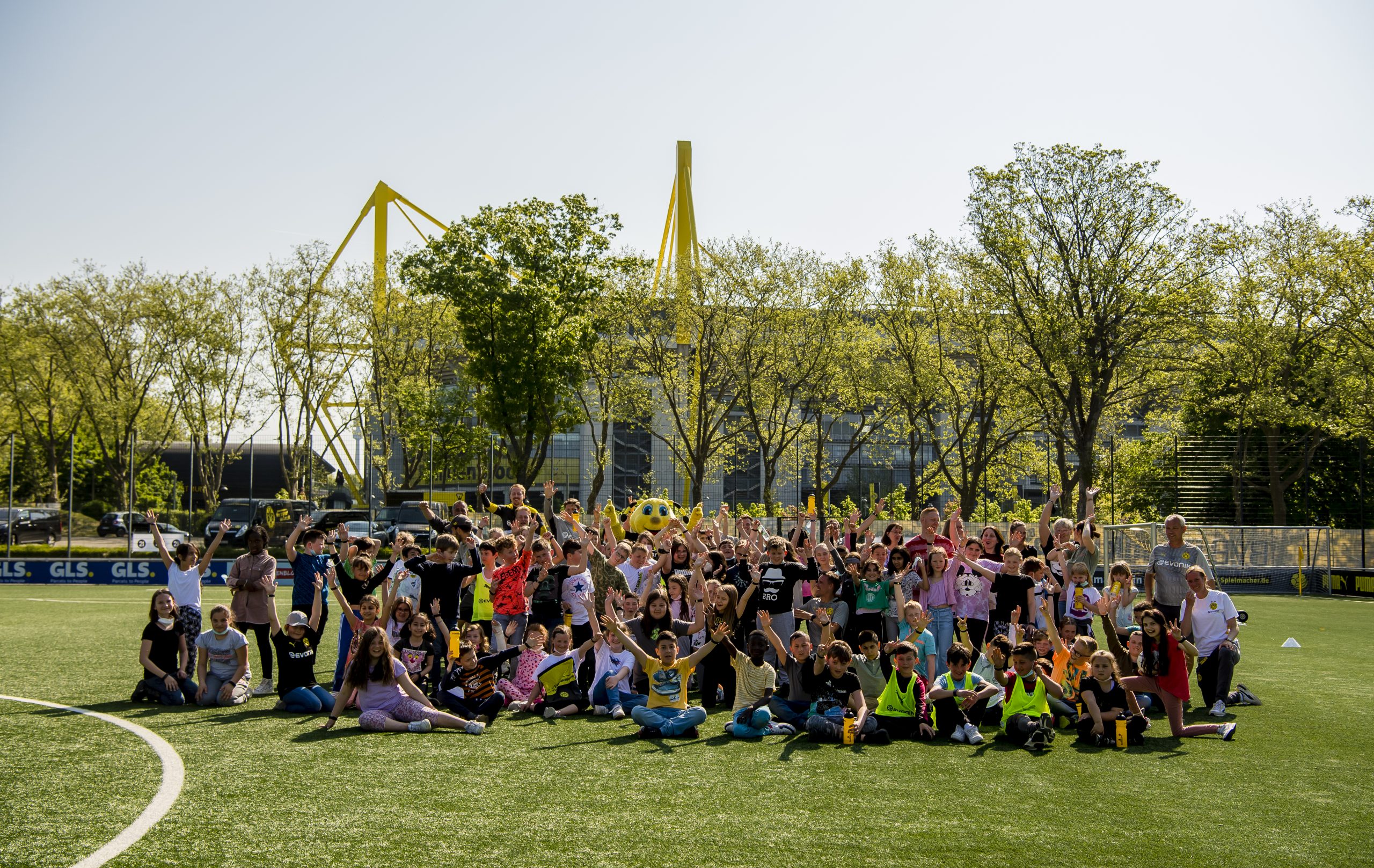 Drei fröhliche Kinder im Fußballtrikot stehen eng umschlungen auf einem Fußballplatz. Links trägt ein Kind eine grüne Weste, die beiden anderen weiße Trikots mit dem Logo von "Indeed" und dem Eintracht Frankfurt Wappen. Im Hintergrund sind andere Spieler und Zuschauer erkennbar.