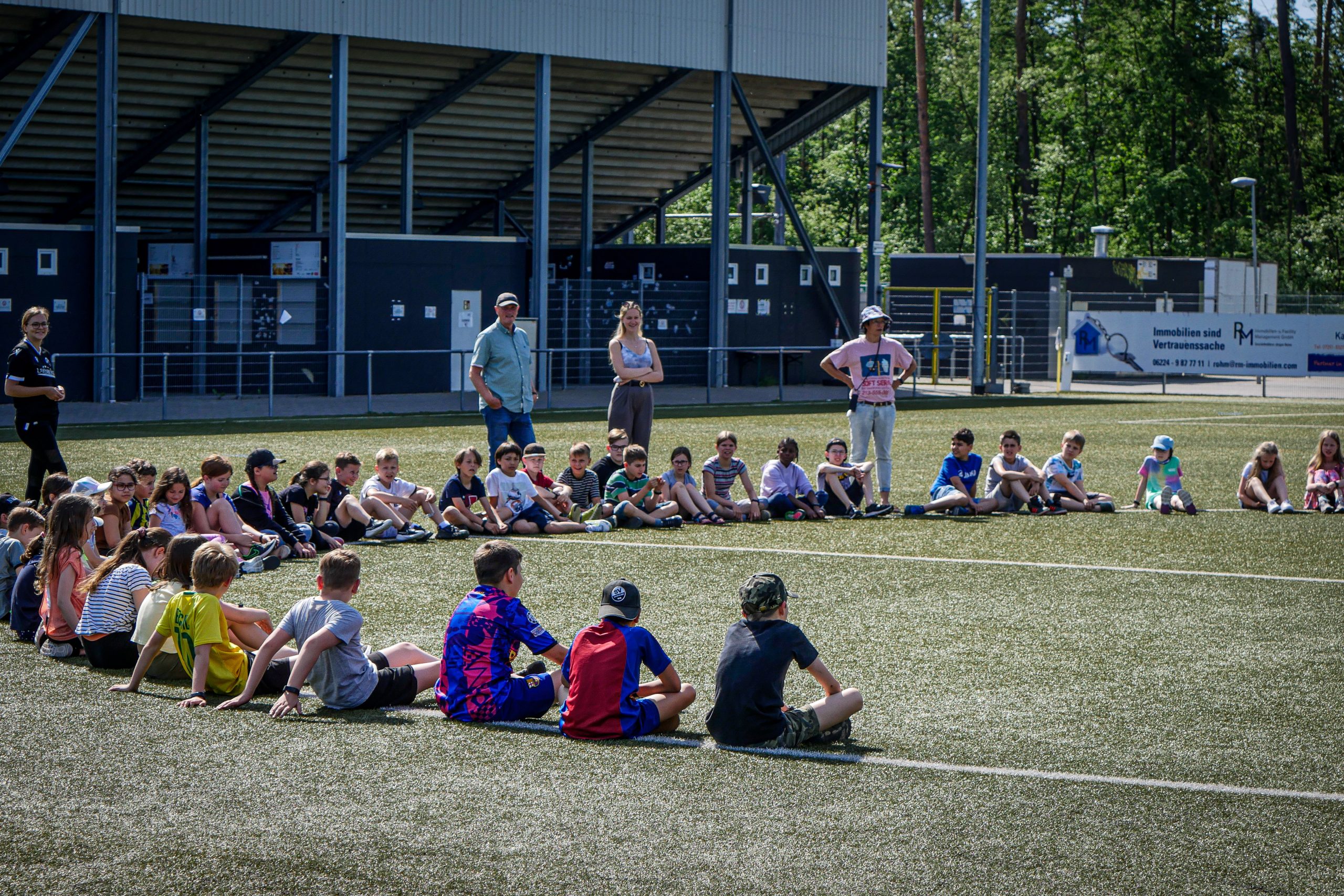 Eine Gruppe von Menschen steht auf einem Fußballplatz vor einem Banner mit der Aufschrift "Willkommen im Fußball - Bundesweites Engagement für junge Geflüchtete". Im Hintergrund ist ein Stadion zu sehen.