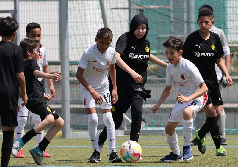 Eine Gruppe von Menschen steht vor einem Banner mit dem Aufdruck "Lernort Stadion". Die Szene zeigt eine Präsentation oder einen Vortrag im Innenraum eines Stadions, vermutlich im Rahmen eines Projekts der DFL Stiftung zur politischen Bildung mit Bezug zu Fußball, Kunst, Kultur und Vereinsgeschichte.