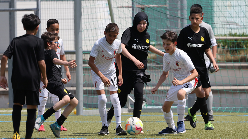 Eine Gruppe von Menschen steht vor einem Banner mit dem Aufdruck "Lernort Stadion". Die Szene zeigt eine Präsentation oder einen Vortrag im Innenraum eines Stadions, vermutlich im Rahmen eines Projekts der DFL Stiftung zur politischen Bildung mit Bezug zu Fußball, Kunst, Kultur und Vereinsgeschichte.