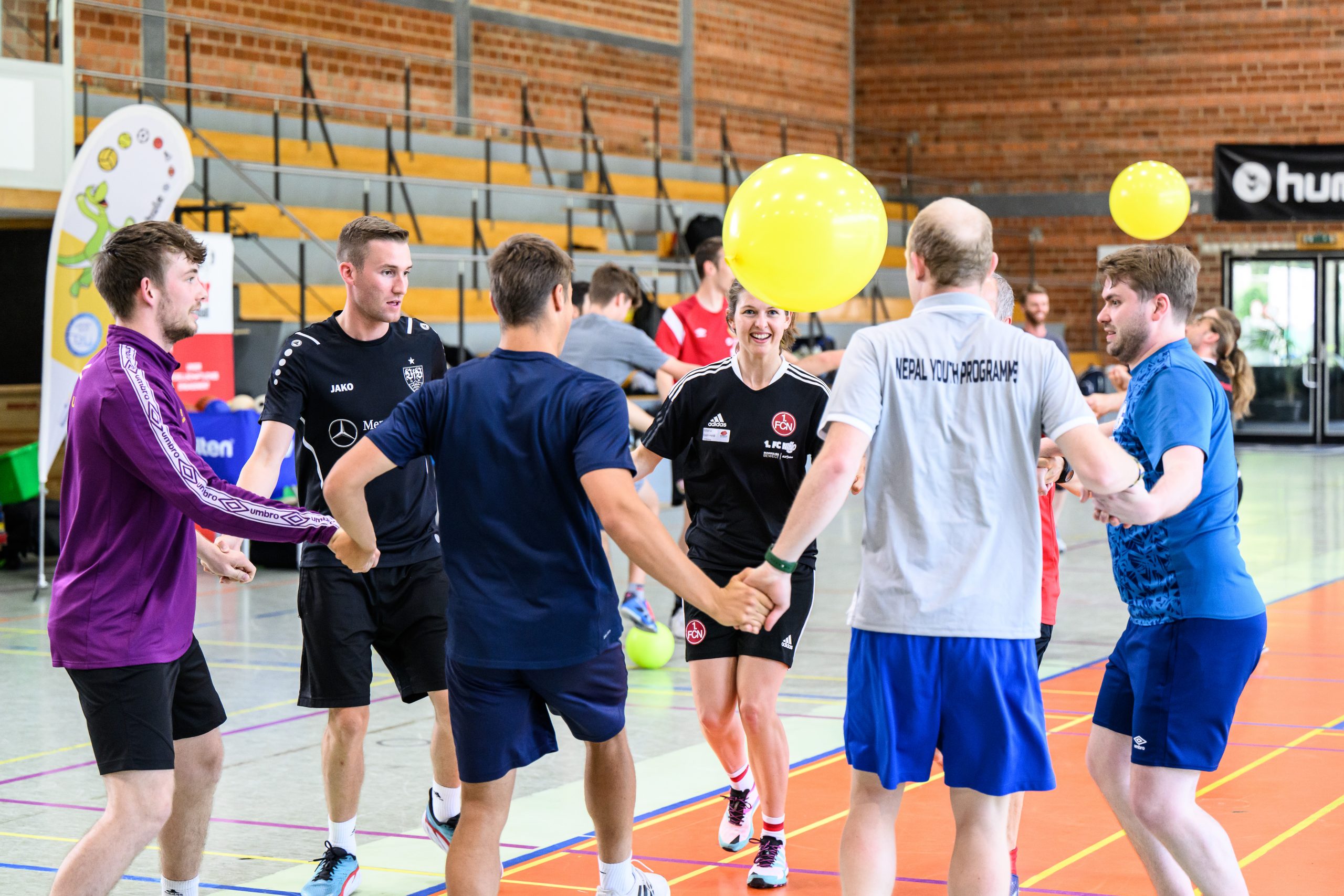 Ein Mädchen mit blonden Haaren springt beim Hochsprung auf einer Bahn. Im Hintergrund sind weitere Kinder und Zuschauer auf einer grünen Wiese zu sehen. Vor dem Mädchen sind Markierungen für die Sprunghöhen aufgestellt.