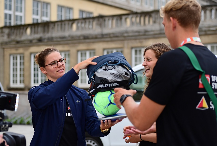 Drei Personen halten verschiedene Sportgeräte hoch.