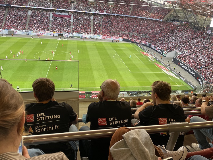 Mit Blick auf das Spielfeld sitzen drei Personen in den neuen T-Shirts der Kampagne #ZukunftBewegen.