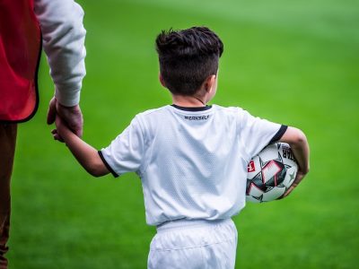 Ein Kind ist mit einem Ball im Arm und einem Erwachsenen an der Hand auf einem Rasenplatz von hinten zu sehen.