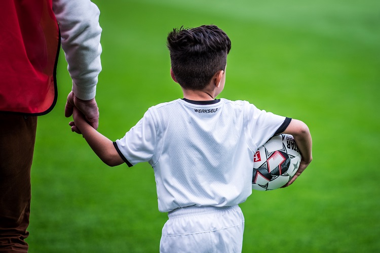 Ein Kind ist mit einem Ball im Arm und einem Erwachsenen an der Hand auf einem Rasenplatz von hinten zu sehen.