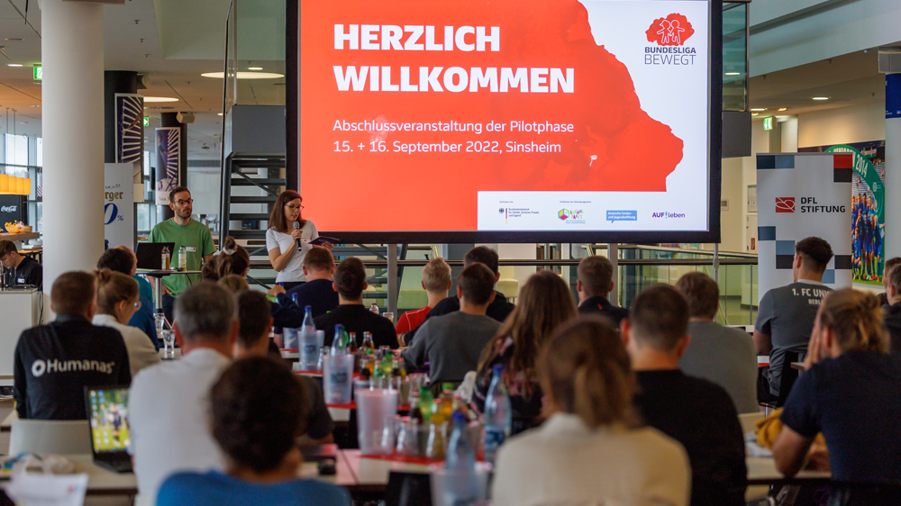 Eine Gruppe von Erwachsenen steht vor einem Banner mit der Aufschrift "Fußball trifft Kultur" und dem Logo der DFL Stiftung. Im Hintergrund ist ein Fußballfeld mit spielenden Kindern erkennbar.