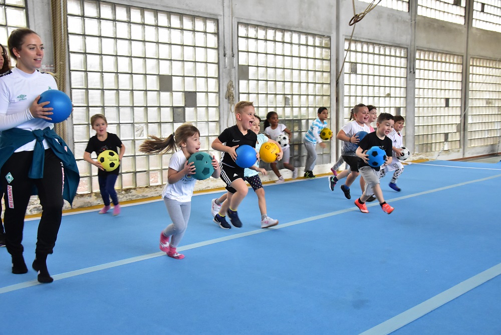 Eine Gruppe junger Menschen steht lächelnd auf einem Fußballfeld. Sie tragen blaue und grüne Trainingswesten und Fußballtrikots. Einige halten Fußbälle oder Wasserflaschen in den Händen. Im Hintergrund ist ein Wohngebäude zu sehen.