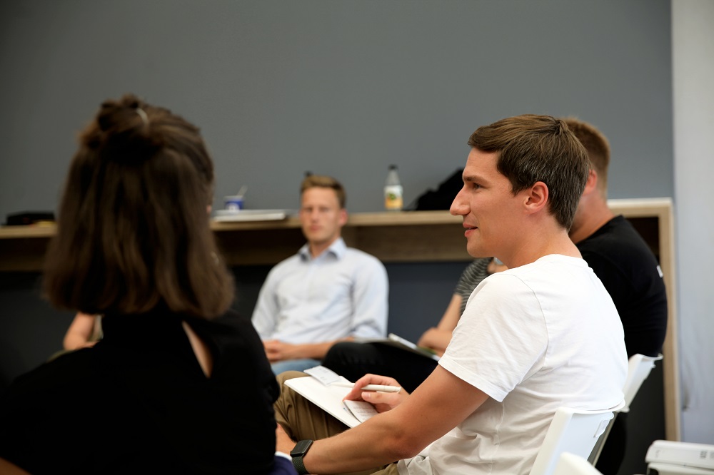 Eine Gruppe Kinder in roten Trikots hält vor einem Fußballstadion ein großes Banner mit der Aufschrift "Fit Vorm Tor!". Im Hintergrund ist ein voll besetztes Stadion zu sehen.