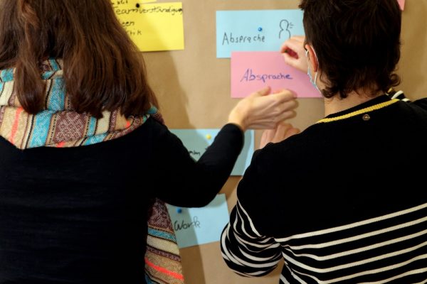 Fußballstadion mit Zuschauern, die die Farben der Regenbogenflagge hochhalten, um ein Zeichen gegen Homophobie zu setzen. Darunter Text: 