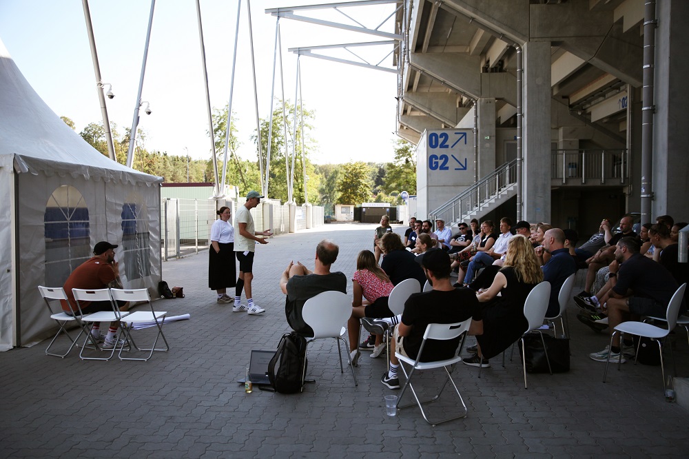 Zwei Fußballspieler sitzen im Kreis mit Teilnehmer*innen eines Workshops. Der Spieler links trägt einen schwarzen Trainingsanzug, der Spieler rechts ein rotes Trikot und hält ein Mikrofon. Im Hintergrund sind weitere Personen und eine Wand mit Plakaten zu sehen.