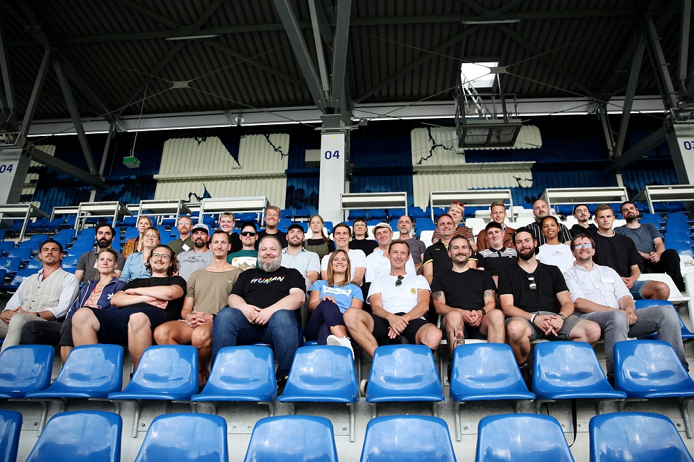"Szene aus dem Café International in Ingolstadt: Im Hintergrund ein Banner mit der Aufschrift "Willkommen im Fußball" und dem Logo der DFL Stiftung. Menschen sitzen an Tischen, einige tragen Fußballtrikots."