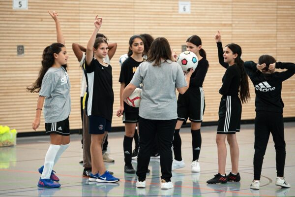 Eine Gruppe von Mädchen steht in einer Turnhalle und spielt Fußball. Einige Mädchen heben ihre Arme, während eine erwachsene Betreuerin mit dem Ball steht. Im Hintergrund ist eine hölzerne Wand zu sehen. Das Bild zeigt eine Szene aus dem Projektbesuch 