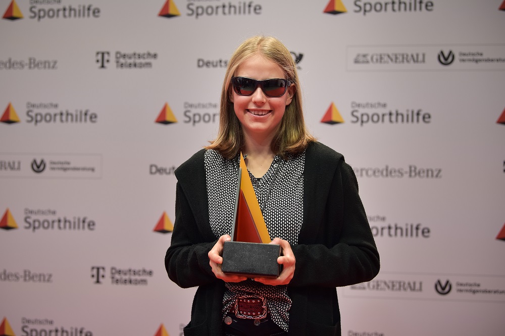 Linn Kazmaier steht vor einer Pressewand mit verschiedenen Logos und hält eine Trophäe in den Händen.