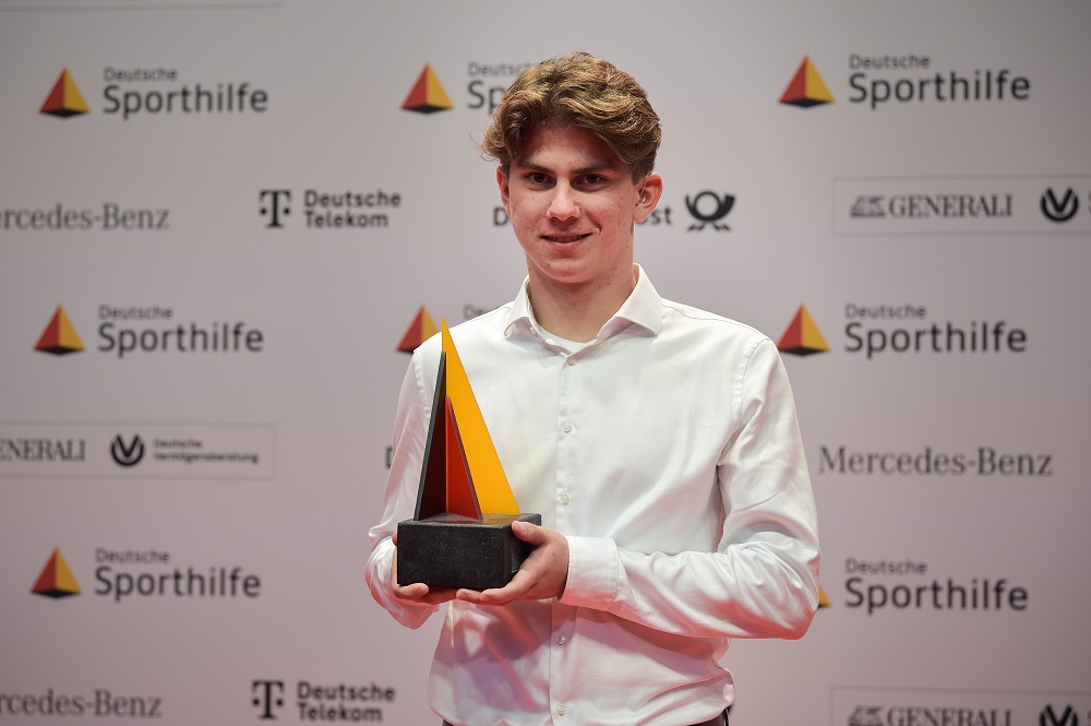 Niklas Müller steht vor einer Pressewand mit verschiedenen Logos und hält eine Trophäe in den Händen.