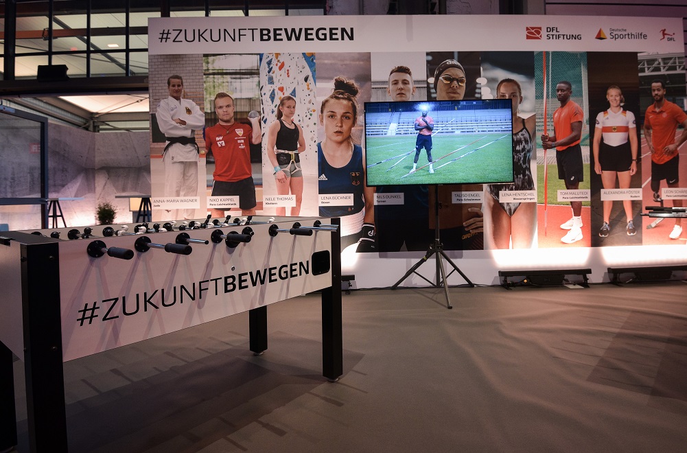 Im Vordergrund steht ein Kicker, im Hintergrund ist eine Wand mit Fotos verschiedener Athlet*innen, dem Hashtag #ZukunftBewegen und einem Fernseher.