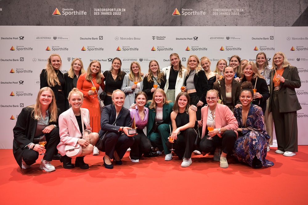 23 junge Frauen stehen bzw. hocken in zwei Reihen vor einer Pressewand.
