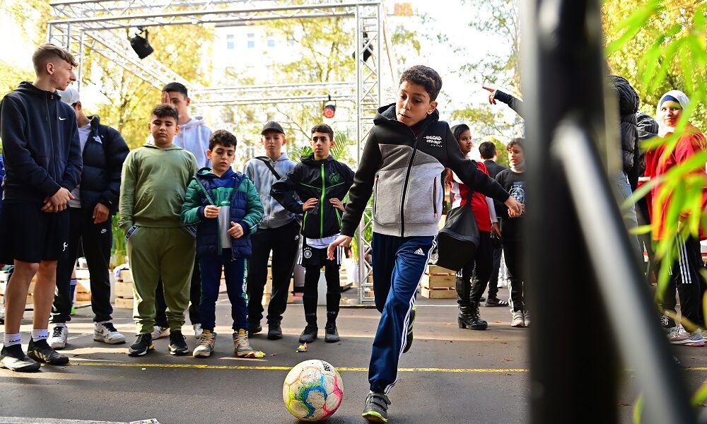 Ein junger Mann wirft einen roten Frisbee auf einem grünen Kunstrasenplatz, umgeben von einem schwarzen Fangnetz.