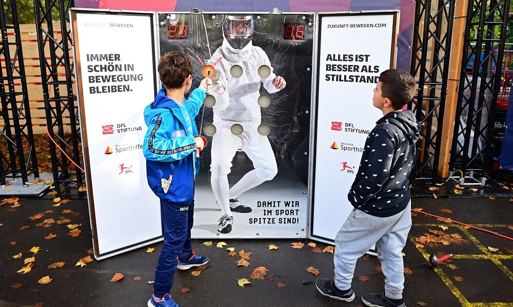 Ein junger Mann mit blonden Haaren und sportlicher Figur steht in einer großen Halle. Er trägt schwarze Sportkleidung und blaue Handschuhe und blickt nach unten. Im Hintergrund sind weitere Trainingsgeräte und die Halle erkennbar.