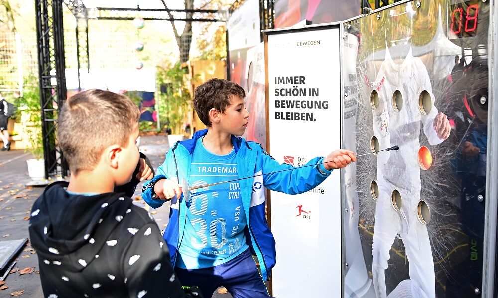 Ein Mann wirft einen Ball in einer Sporthalle, während ein Kamerateam ihn filmt. Im Hintergrund sind weitere Personen und Sportgeräte zu sehen.
