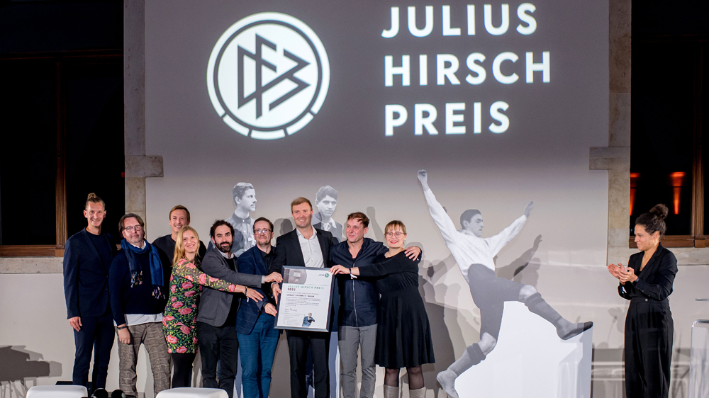 Auf einer Bühne stehen neun Personen. Fünf von ihnen halten eine Urkunde in der Hand. Im Hintergrund steht an die Wand projiziert: Julius Hirsch Preis.