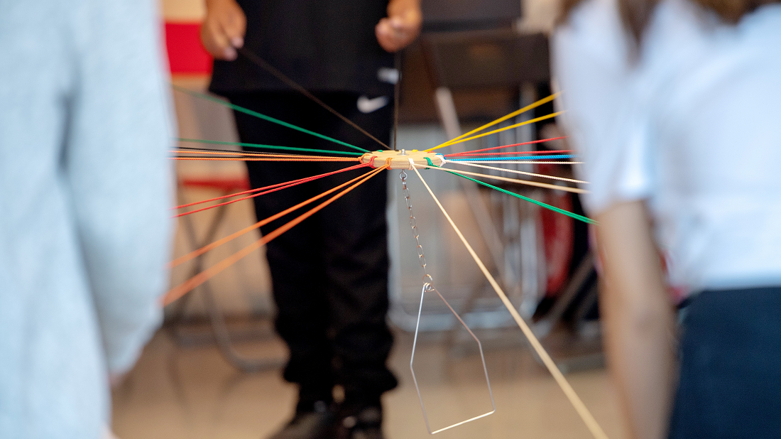 Ein komplexes Netz aus bunten Fäden, die an einem zentralen Knotenpunkt zusammenlaufen, wird von einer Person gehalten. Im Hintergrund sind verschwommene Personen und ein Raum zu erkennen.