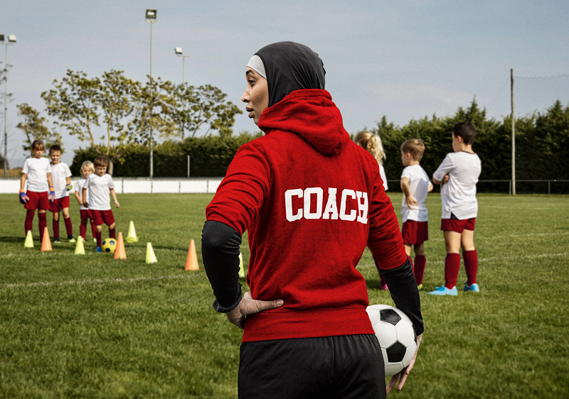Eine Trainerin hält einen Ball in den Händen und steht auf dem Trainingsplatz, während Kinder eine Fußballübung durchführen.