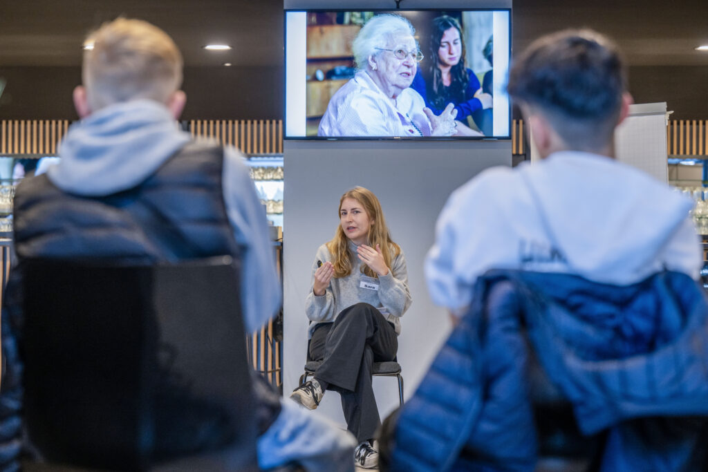 Sara Elkmann vom ZWEITZEUGEN e.V. hält einen Workshop vor einer Gruppe Jugendlicher. Im Hintergrund ist auf einem Bildschirm eine Holocaust-Überlebende zu sehen, die ihre Geschichte erzählt.