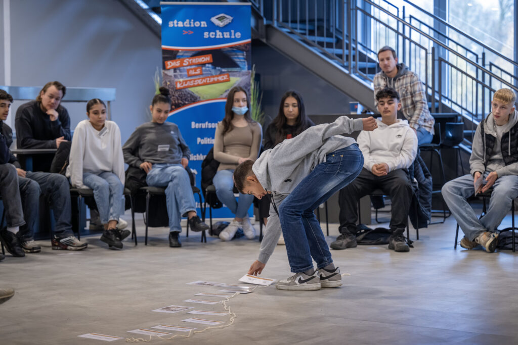 Ein Jugendlicher beugt sich im Rahmen eines Workshops in der Stadionschule Bielefeld vor einer Gruppe von Sitzenden vor und deutet mit dem Finger auf den Boden. Im Hintergrund ist eine Treppe und ein Banner mit dem Aufdruck "Stadion | Schule" zu sehen. Der Workshop beschäftigt sich mit der Geschichte der nationalsozialistischen Judenverfolgung und der aktiven Auseinandersetzung mit Antisemitismus.