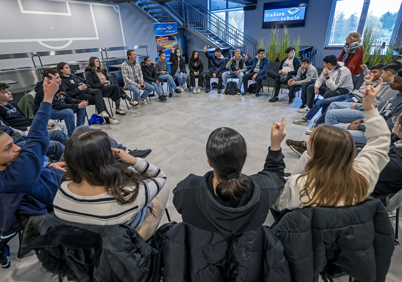 Eine Gruppe Jugendlicher sitzt im Kreis auf Stühlen in einer hellen, modernen Aula und hält die Hände gehoben. Einige blicken erwartungsvoll in die Runde. Im Hintergrund sind eine Treppe und Plakate zu sehen.