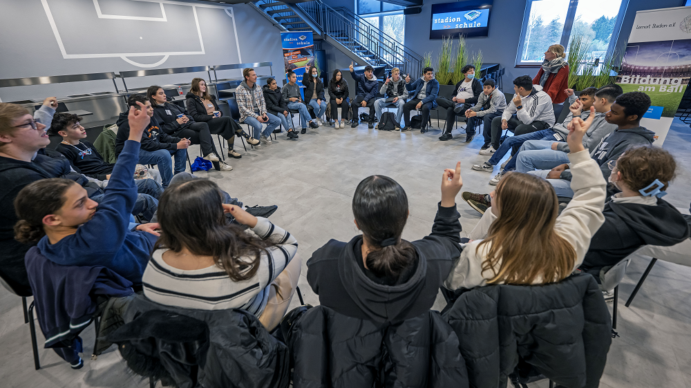Eine Gruppe Jugendlicher sitzt im Kreis auf Stühlen in einer hellen, modernen Aula und hält die Hände gehoben. Einige blicken erwartungsvoll in die Runde. Im Hintergrund sind eine Treppe und Plakate zu sehen.