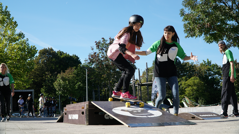Ein Mädchen übt das Skateboardfahren, eine Betreuerin hilft ihr dabei.