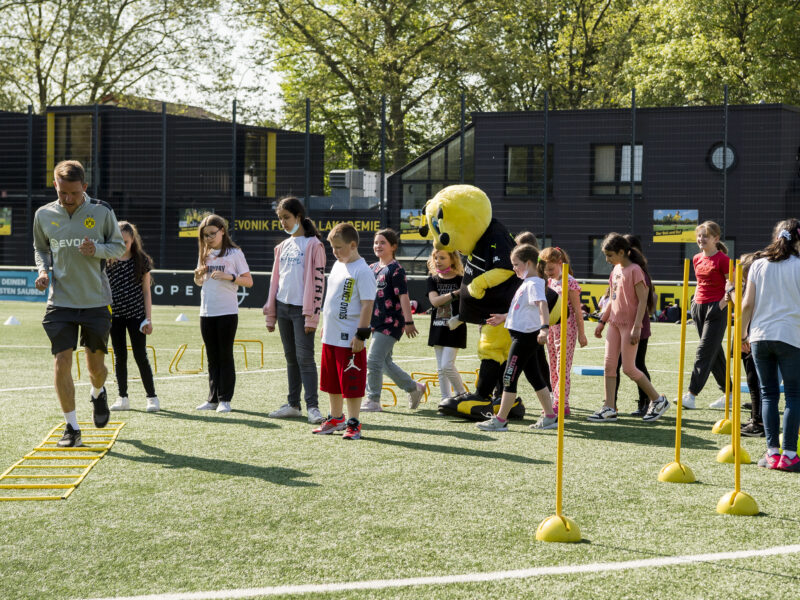 Eine Gruppe von Kindern und Jugendlichen nimmt an einem Sportevent auf dem Fußballplatz der Borussia Dortmund Akademie teil. Ein Maskottchen der Borussia Dortmund begleitet die Gruppe. Im Hintergrund ist ein Gebäude der Akademie zu sehen.
