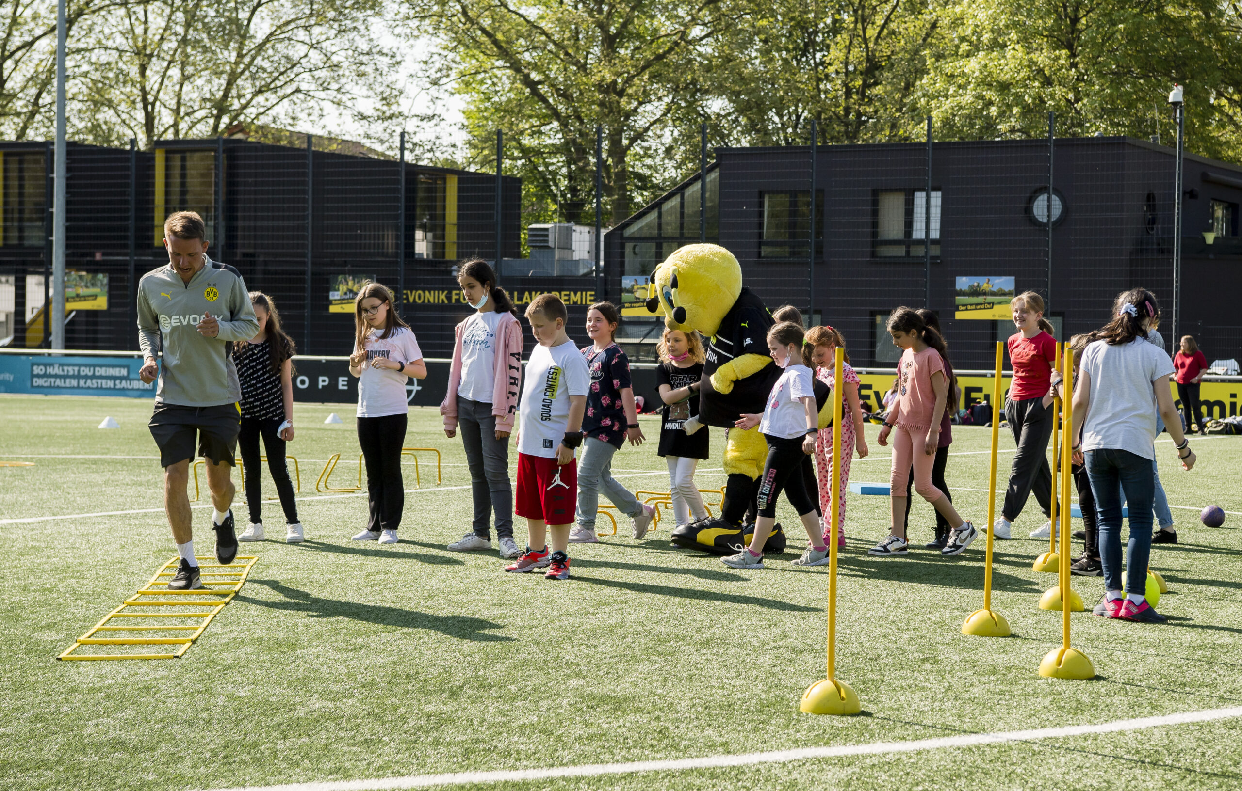 Eine Gruppe von Kindern und Jugendlichen nimmt an einem Sportevent auf dem Fußballplatz der Borussia Dortmund Akademie teil. Ein Maskottchen der Borussia Dortmund begleitet die Gruppe. Im Hintergrund ist ein Gebäude der Akademie zu sehen.