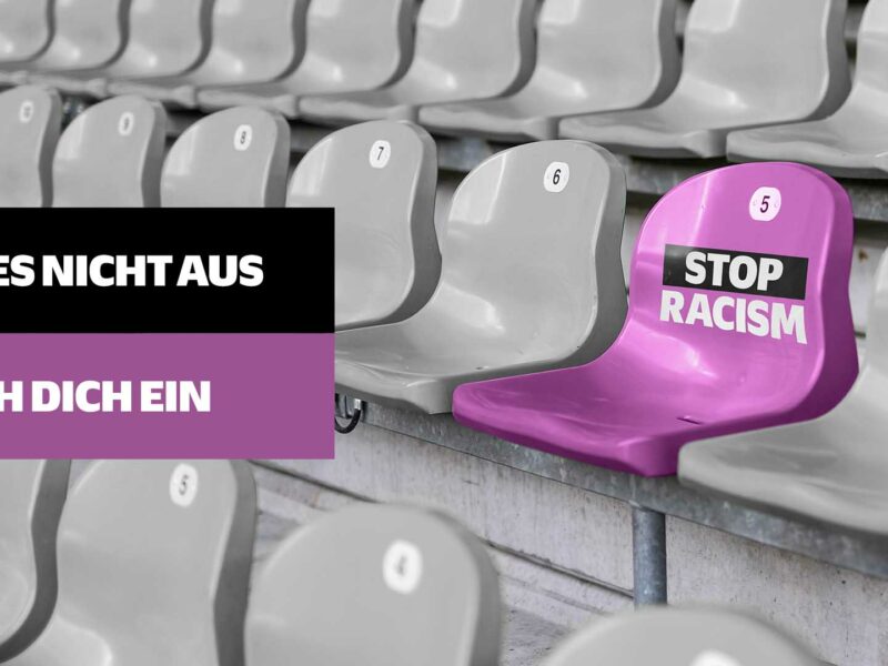 Ein leerer violetter Stadionstuhl mit dem Aufdruck "STOP RACISM" steht zwischen grauen Stadionstühlen. Im Vordergrund ein violettes Banner mit der Aufschrift "SITZ ES NICHT AUS - MISCH DICH EIN". Das Bild dient als Aufruf gegen Rassismus im Fußball.