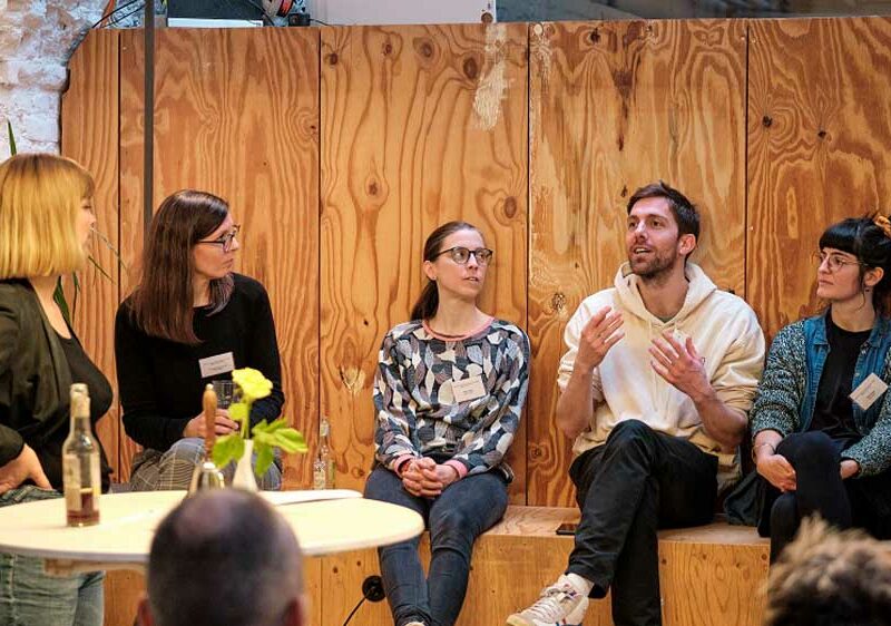 Gruppe von Menschen sitzt in einem Workshop-Setting auf einer Holzbank und diskutiert. Ein Mann gestikuliert dabei lebhaft, während die anderen ihm aufmerksam zuhören. Im Vordergrund steht ein Tisch mit Wasserflaschen und Blumen.
