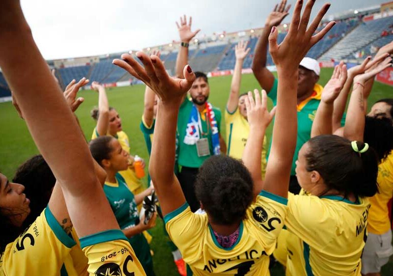 Eine Gruppe junger Menschen steht auf einem Fußballfeld im Kreis. Die jungen Menschen heben ihre Arme in die Luft.
