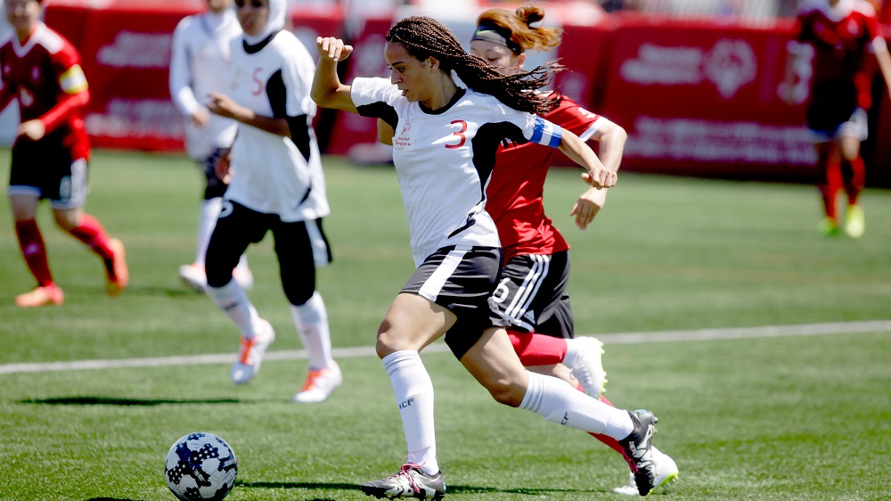 Eine Fußballspielerin mit langen, dunklen Haaren dribbelt auf einem grünen Rasen einen Fußball. Im Hintergrund sind weitere Spielerinnen in roten und weißen Trikots zu sehen.