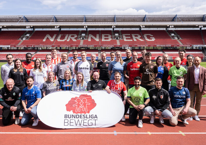 Eine große Gruppe von Menschen steht auf der roten Tartanbahn eines Fußballstadions vor der Zuschauertribüne. Viele tragen T-Shirts von Fußballvereinen oder Sportbekleidung. In der Mitte hält eine Frau ein Banner mit der Aufschrift "BUNDESLIGA BEWEGT" mit einem stilisierten Logo aus mehreren Figuren. Im Hintergrund ist die leere Zuschauertribüne des Stadions zu sehen.