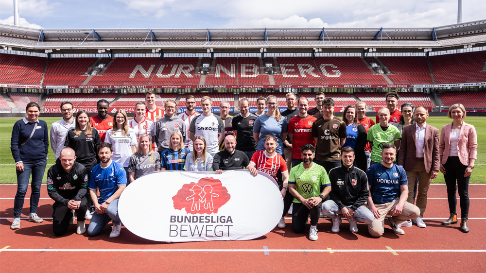 Eine große Gruppe von Menschen steht auf der roten Tartanbahn eines Fußballstadions vor der Zuschauertribüne. Viele tragen T-Shirts von Fußballvereinen oder Sportbekleidung. In der Mitte hält eine Frau ein Banner mit der Aufschrift "BUNDESLIGA BEWEGT" mit einem stilisierten Logo aus mehreren Figuren. Im Hintergrund ist die leere Zuschauertribüne des Stadions zu sehen.