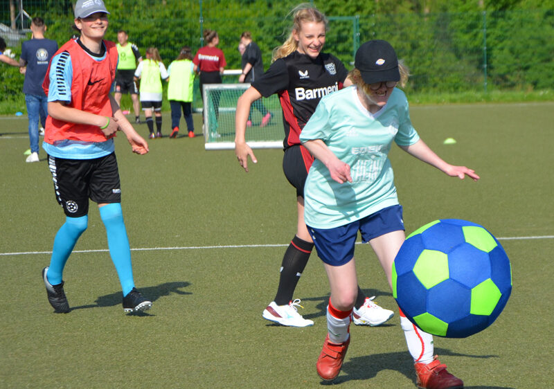 Eine junge Frau mit Cap und hellgrünem T-Shirt schießt einen großen, blauen Fußball auf einem grünen Kunstrasenplatz. Im Hintergrund sind weitere Teilnehmende einer Trainingsgruppe zu sehen.
