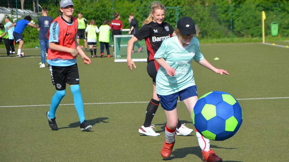 Eine junge Frau mit Cap und hellgrünem T-Shirt schießt einen großen, blauen Fußball auf einem grünen Kunstrasenplatz. Im Hintergrund sind weitere Teilnehmende einer Trainingsgruppe zu sehen.