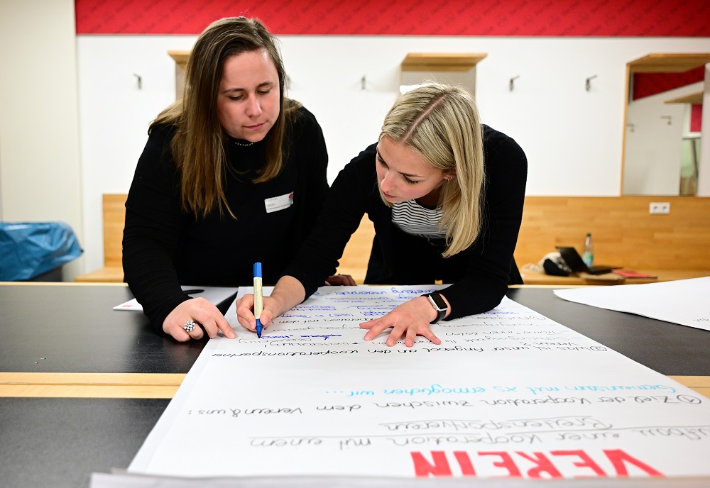Zwei Frauen stehen an einem großen Flipchart und schreiben mit Stiften darauf. Im Hintergrund sind weitere Flipcharts und eine helle Raumgestaltung zu sehen. Es handelt sich um eine Netzwerkveranstaltung der DFL Stiftung "Bundesliga bewegt".