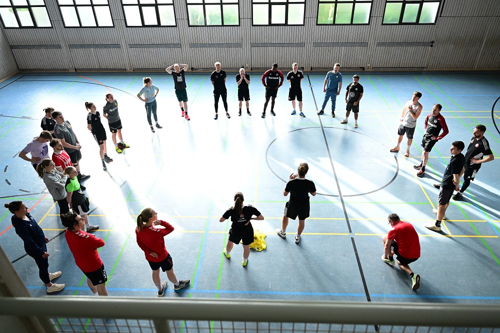 Fußballspieler und Trainer stehen im Kreis in einer Turnhalle, vermutlich während einer Netzwerkveranstaltung der DFL Stiftung "Bundesliga bewegt" in Nürnberg am 27. April 2023.