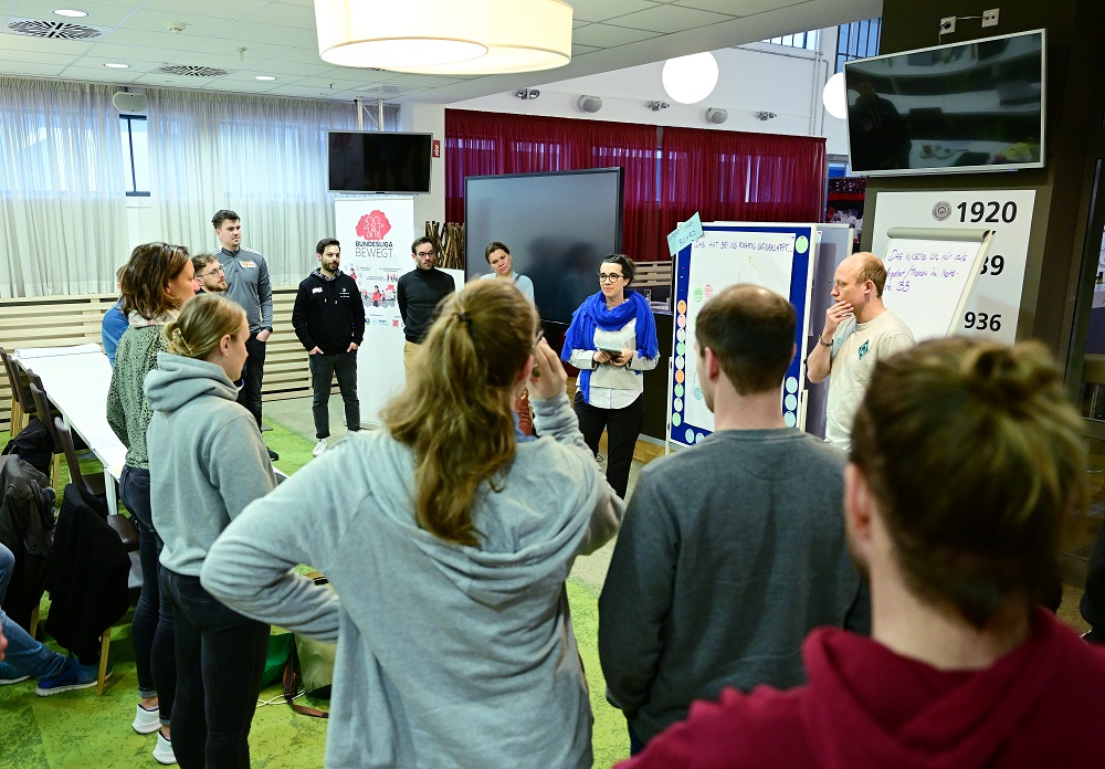 Eine Gruppe von Menschen steht in einem hellen Raum in einem Kreis zusammen und diskutiert. Im Hintergrund sind ein roter Vorhang und Plakate mit dem Logo "Bundesliga bewegt" zu sehen. Das Bild zeigt eine Netzwerkveranstaltung der DFL Stiftung in Nürnberg am 28.04.2023.