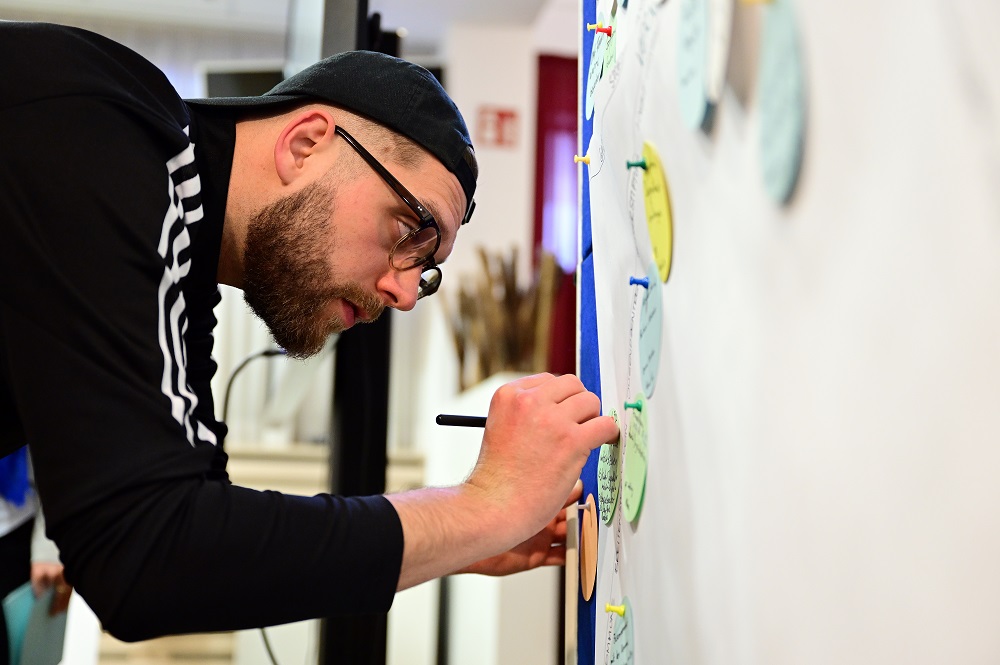 Ein Mann mit Bart und dunkler Kappe befestigt mit einem Stift eine Postkarte an einer Pinnwand mit vielen weiteren Postkarten. Im Hintergrund sind weitere Pinnwände zu sehen. Das Bild stammt von der Netzwerkveranstaltung "Bundesliga bewegt" der DFL Stiftung in Nürnberg am 28.04.2023.