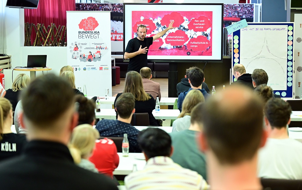 Ein Vortragender steht vor einer Gruppe von Teilnehmenden bei einer Netzwerkveranstaltung der DFL Stiftung mit dem Titel "Bundesliga bewegt". Auf der Präsentation im Hintergrund sind verschiedene Schwerpunkte wie "Fan- und Zuschauer-Service" und "Fußballschule" zu sehen.