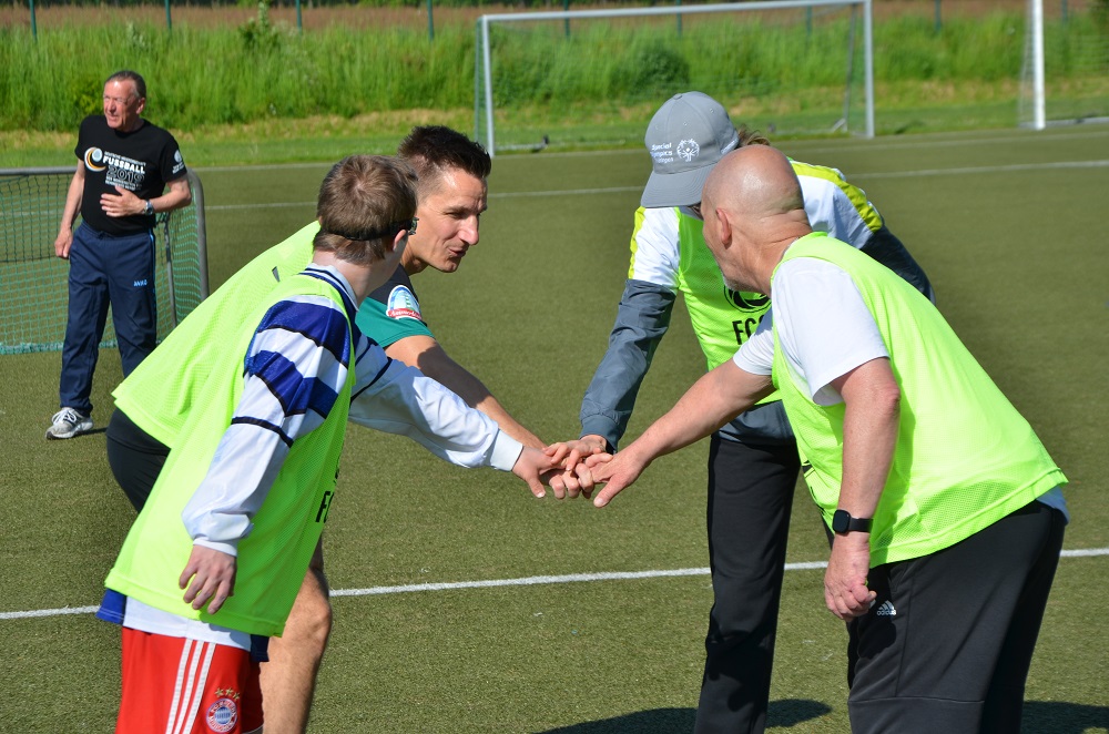 Zwei Männer in Trainingsanzügen und leuchtenden Westen berühren sich mit den Händen und besprechen etwas auf einem Fußballplatz, während ein dritter Mann im Hintergrund zuschaut.