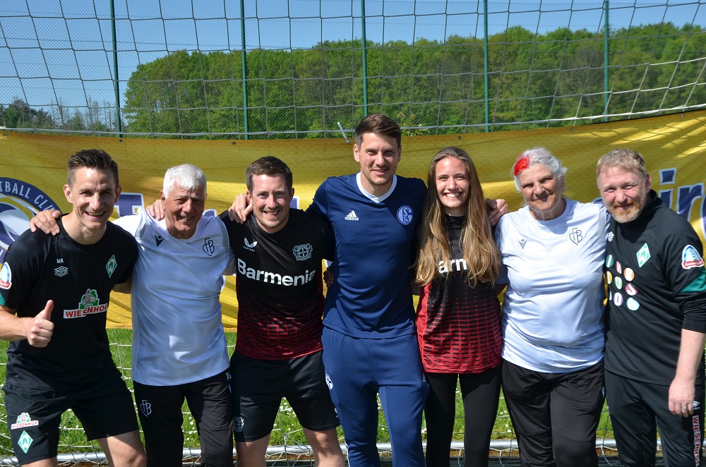 Eine Gruppe von sieben Personen steht lächelnd auf einem Fußballplatz. Sie tragen Sportkleidung verschiedener Fußballvereine und wirken wie Trainer oder Betreuer. Im Hintergrund ist ein grüner Rasen und ein Fußballtor zu sehen.