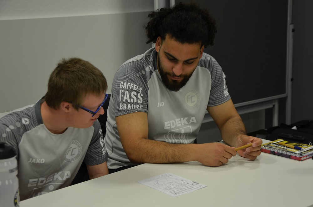 Zwei junge Männer sitzen an einem Tisch und studieren gemeinsam Unterlagen. Beide tragen Trikots mit dem Logo der DFL Stiftung und dem Schriftzug EDEKA. Einer trägt eine Brille und zeigt auf die Papiere, während der andere konzentriert darauf blickt.